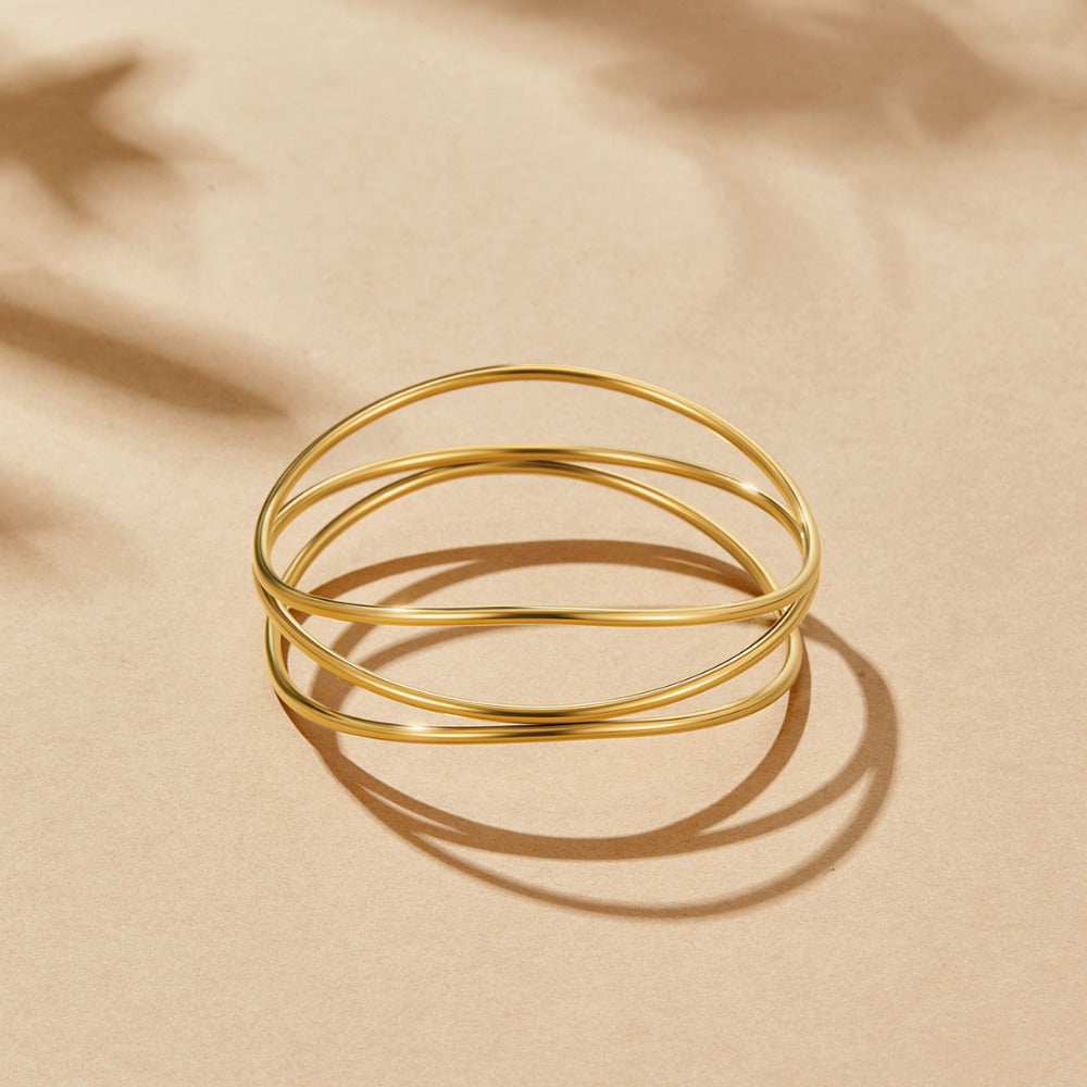 Sabrina Gold Layered Bangle, three thin gold bangles interlaced, lying flat on sand-coloured