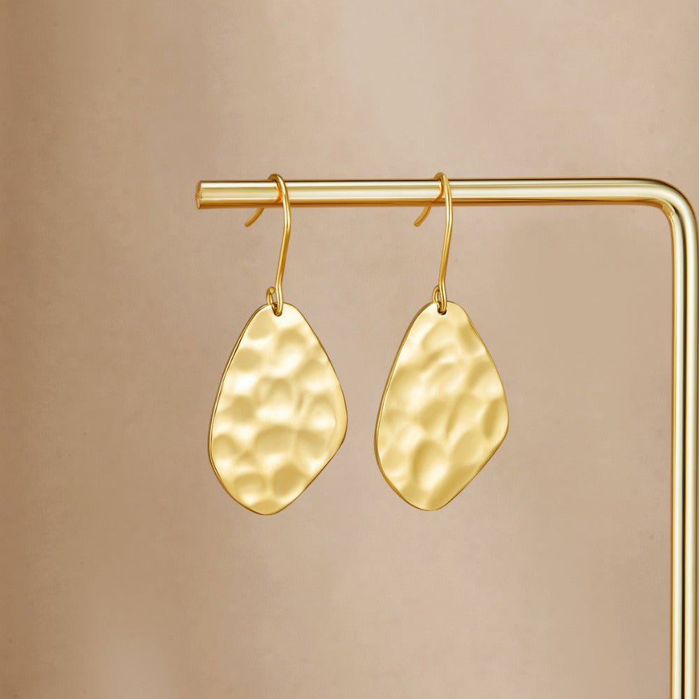 Amaya Hammered Gold Drop Earrings, hanging on a gold display bar against a beige backdrop