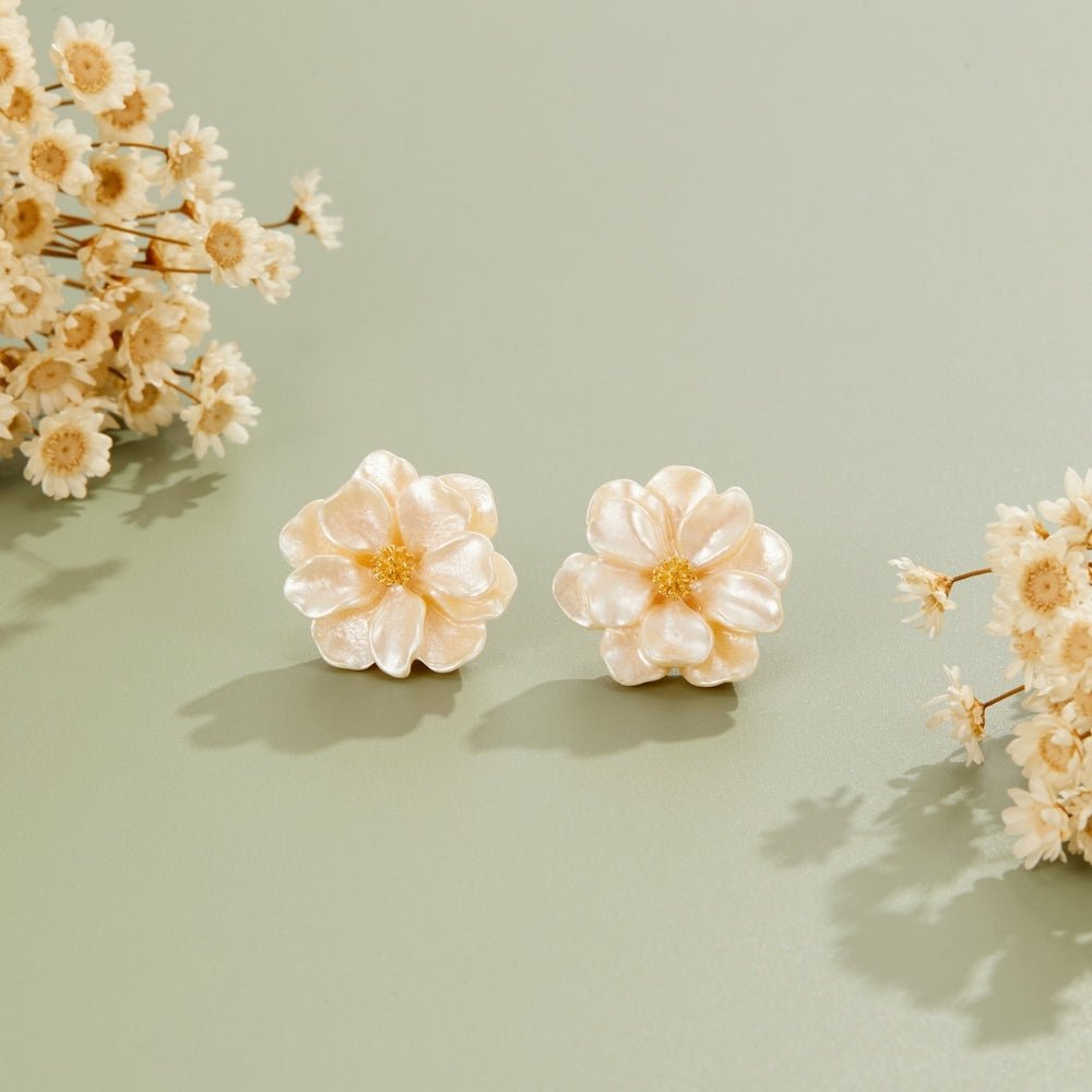 Camelia Bloom Earrings, a pair of pearly cream flower stud earrings on sage green, beside