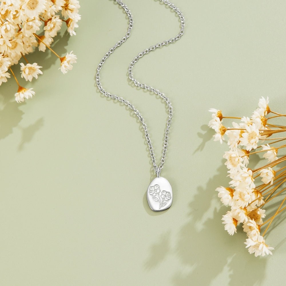 Engraved Wildflower Silver Necklace, laid flat on green surface with dried white flowers, oval