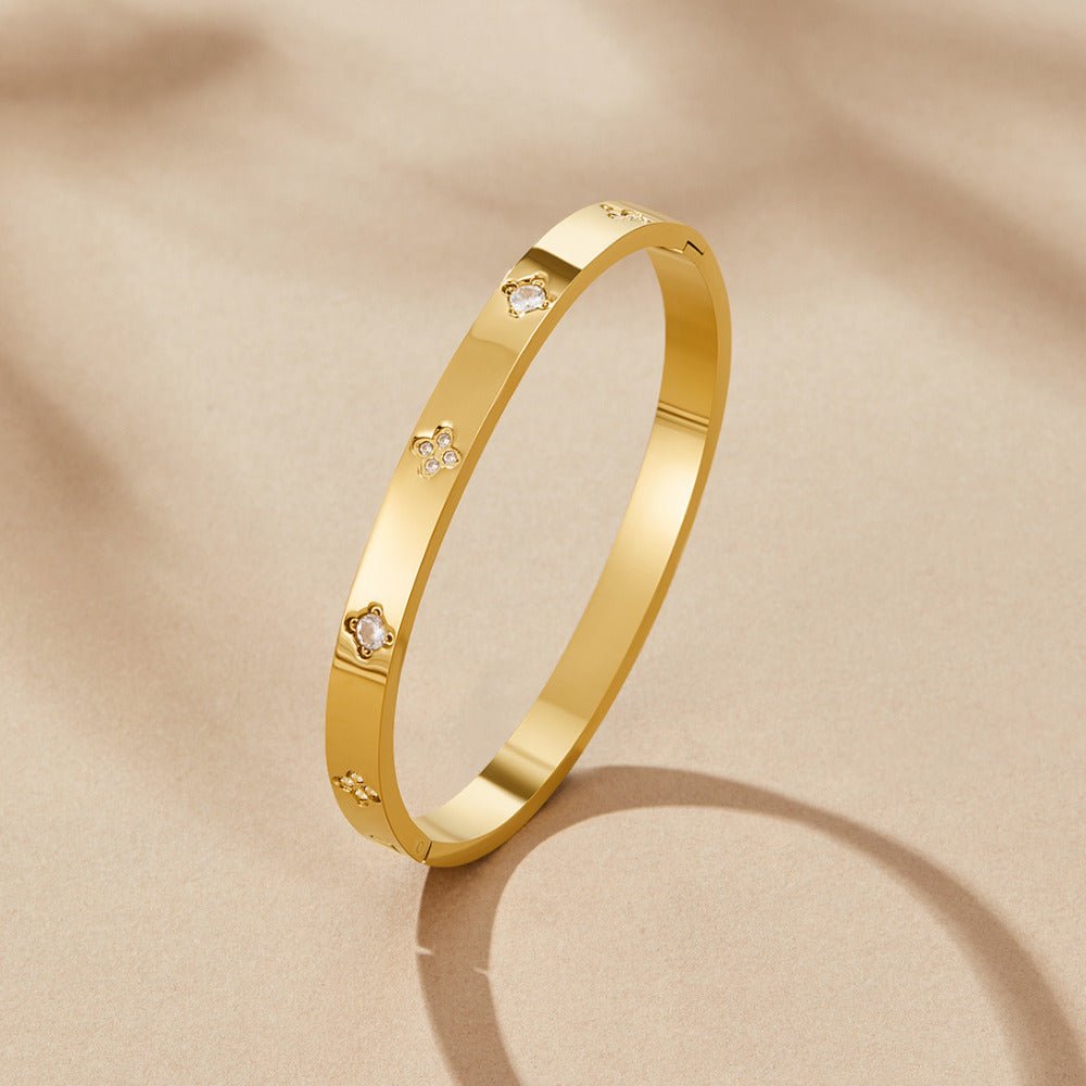 Gold Clover Bangle, standing upright on a beige surface, with clover motifs set with clear stones
