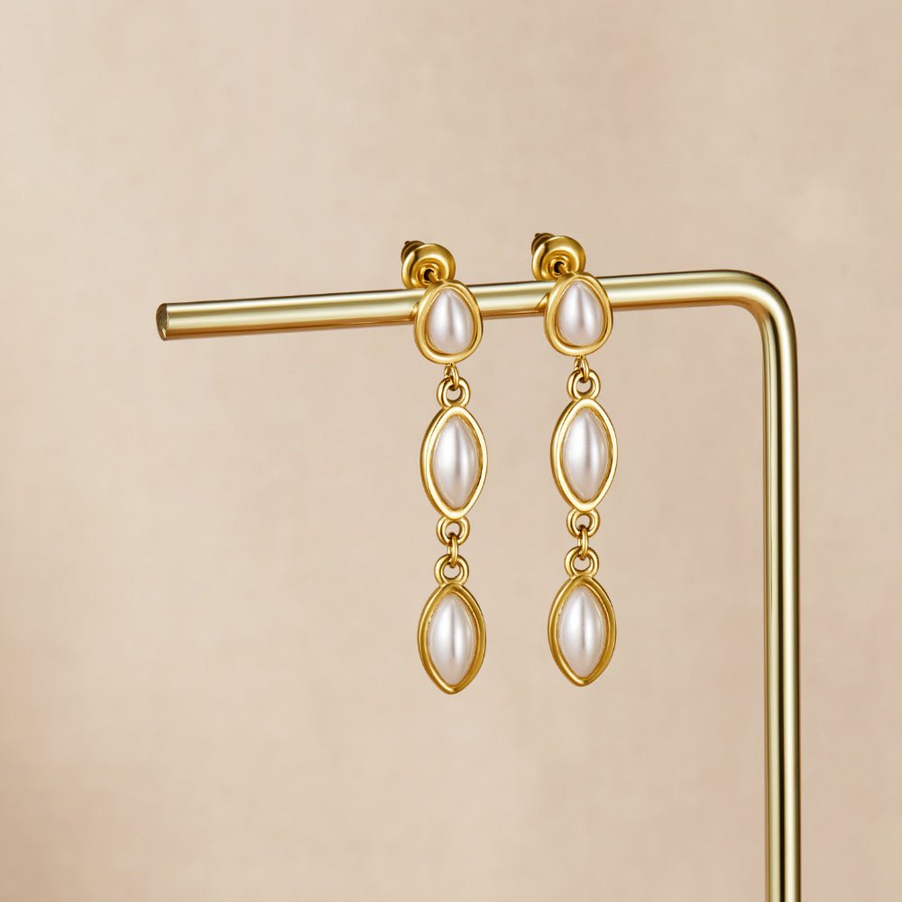 Gold Serena Pearl Drop Earrings, hanging on a gold display stand against a beige backdrop