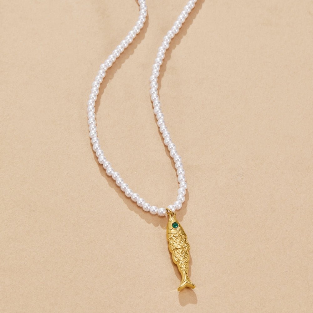Golden Fish Pearl Necklace, pearl necklace laid flat on sand with gold fish pendant and green