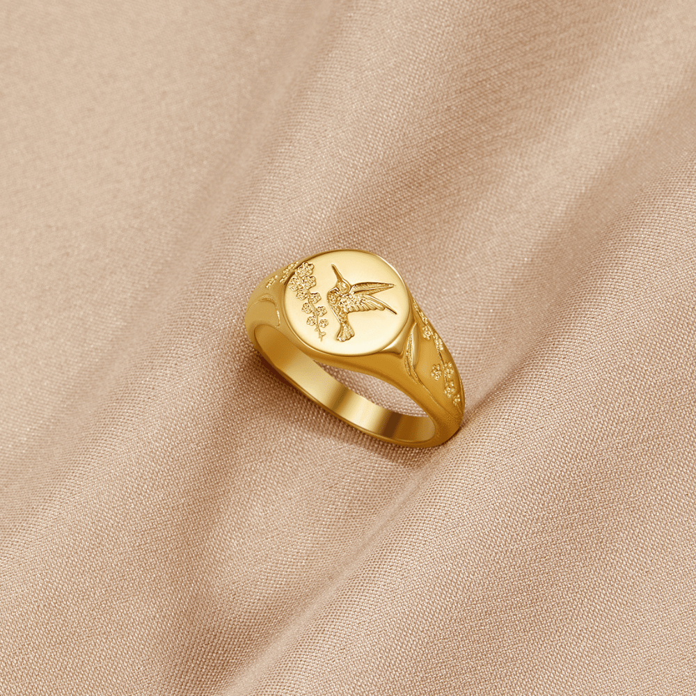Golden Hummingbird Signet Ring, gold signet ring lying on a beige fabric, engraved hummingbird and sprigs