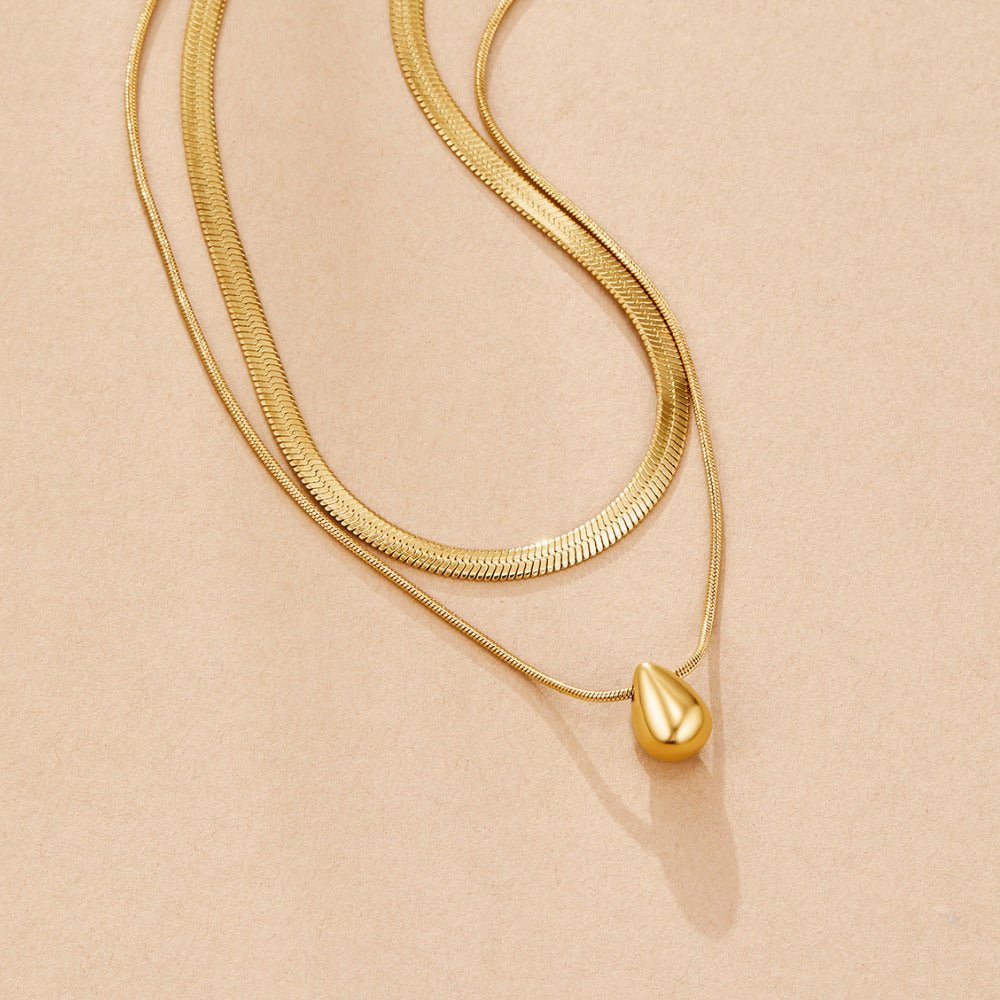 Layered Bella Teardrop Necklace, two gold chains laid flat on a beige surface, teardrop