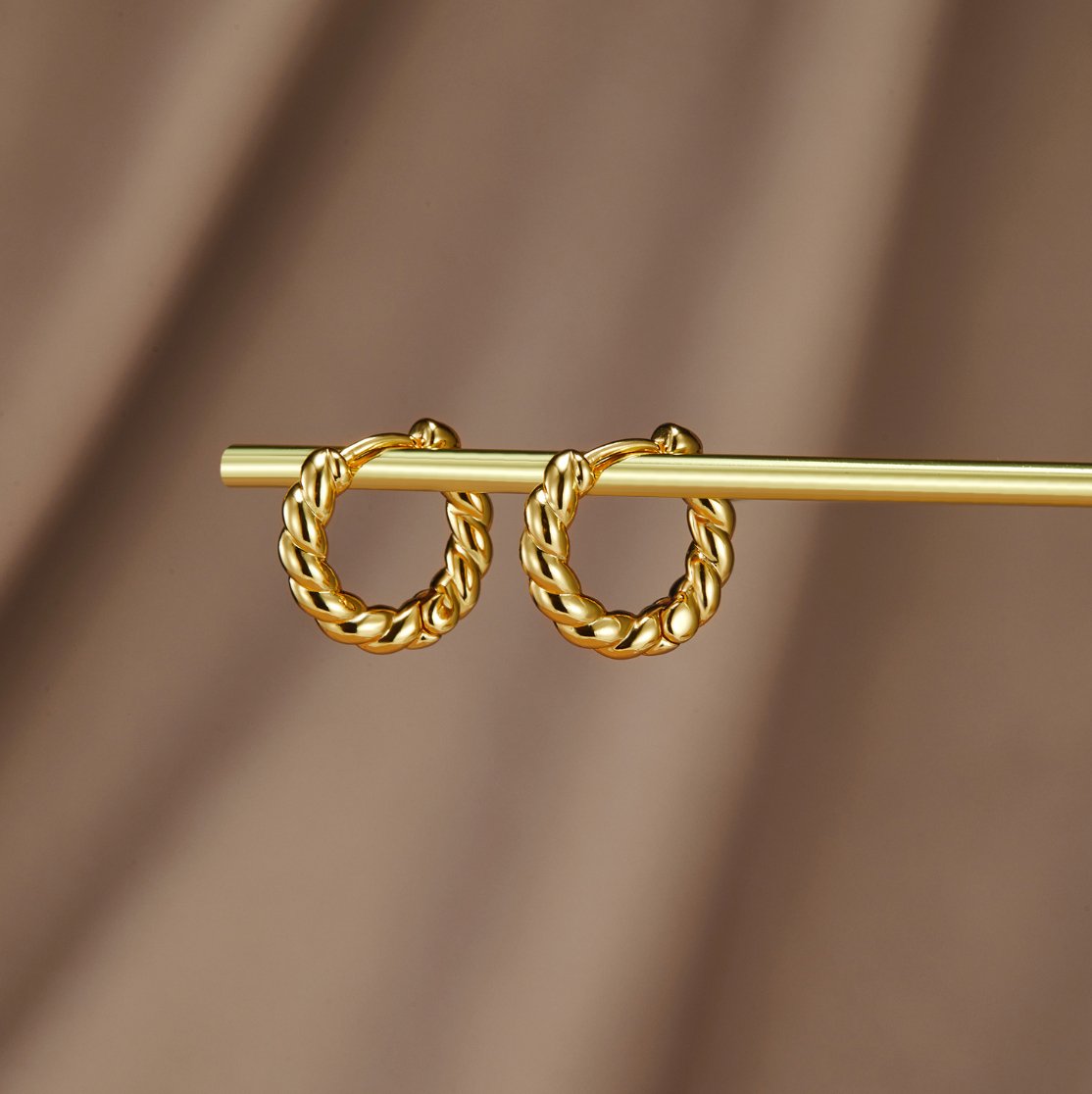 Layla Gold Twist Earrings, pair of twisted gold hoops hanging on a gold rod against brown fabric