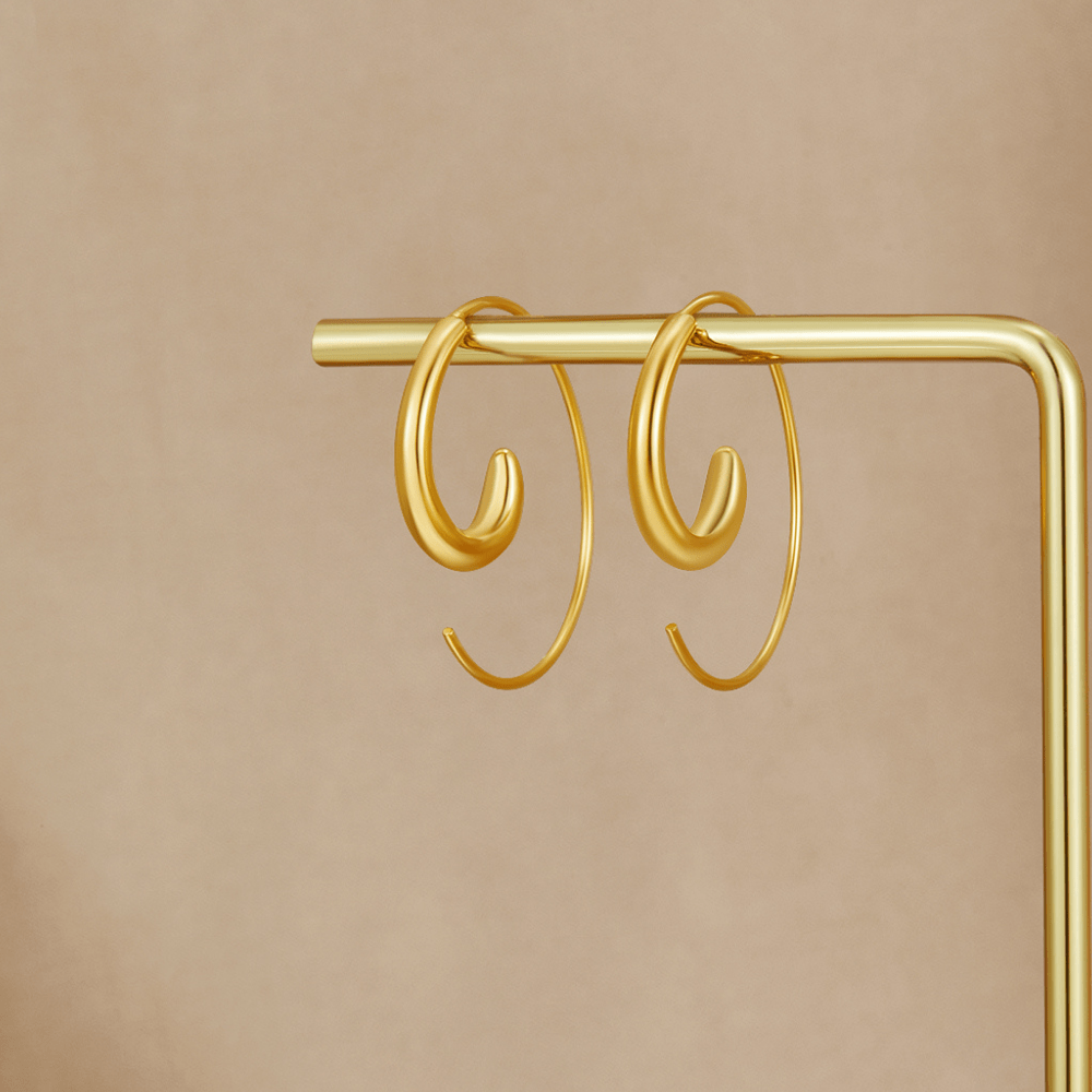 Luca Spiral Hoop Earrings, hanging on a gold display bar against a beige backdrop
