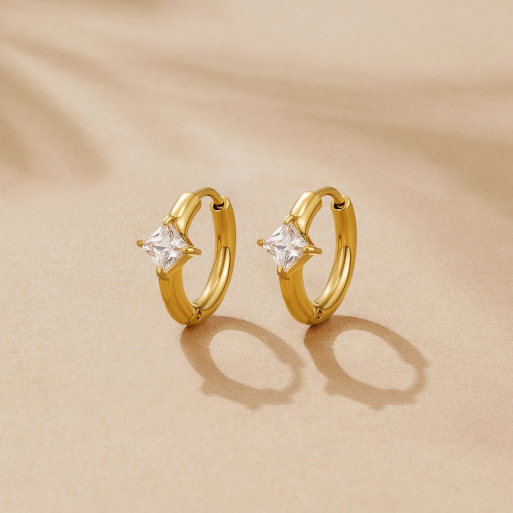 Roselle Crystal Huggies, pair of gold hoops with square clear stones, resting on a beige surface