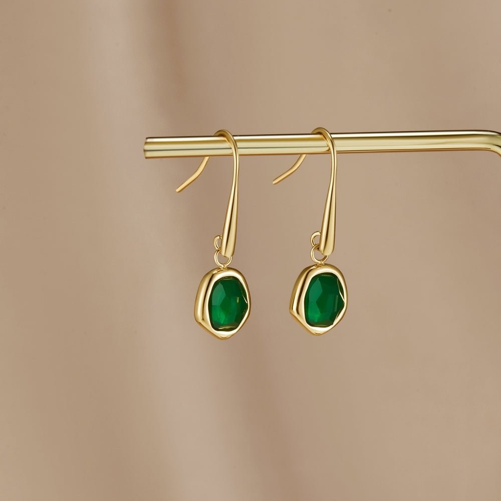 Vintage Emerald Drop Earrings, hanging from a gold bar on a beige backdrop, green stones visible