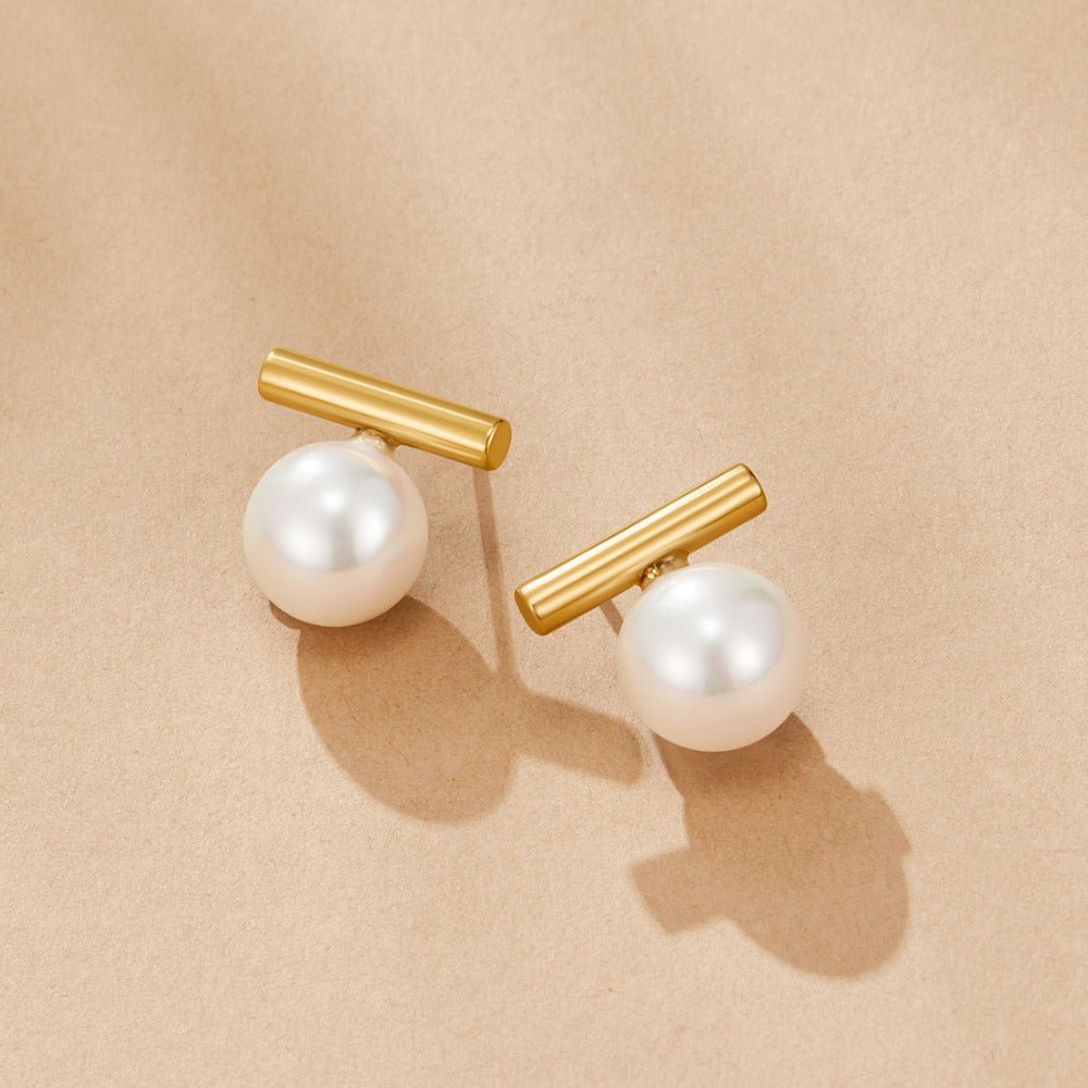 Yuna Pearl Stud Earrings, a pair of gold bar earrings with white pearls on beige paper