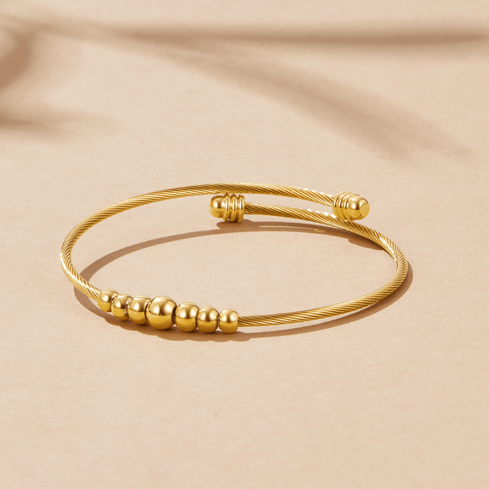 Yuri Gold Bead Bangle, open twisted gold bangle with graduated beads, on a beige surface