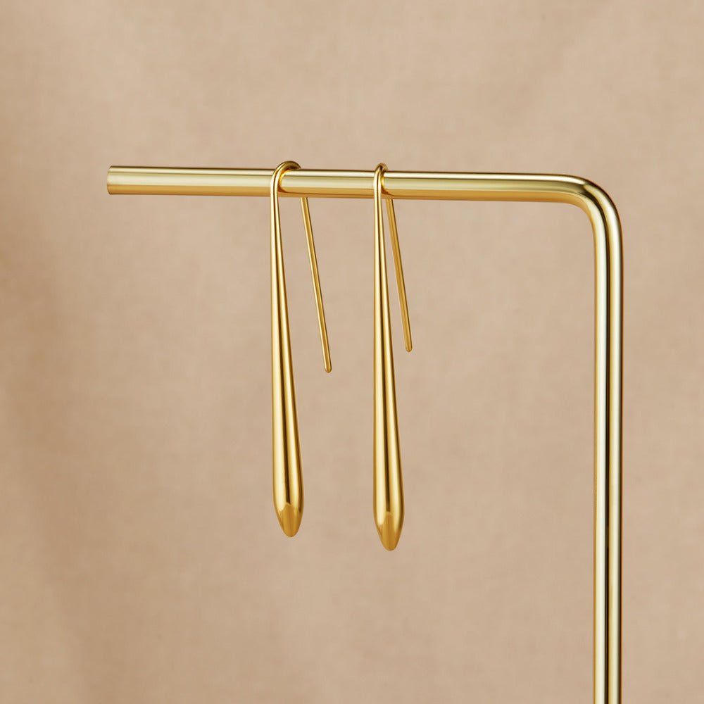 Zia Gold Drop Earrings, hanging on a gold display bar against a beige backdrop