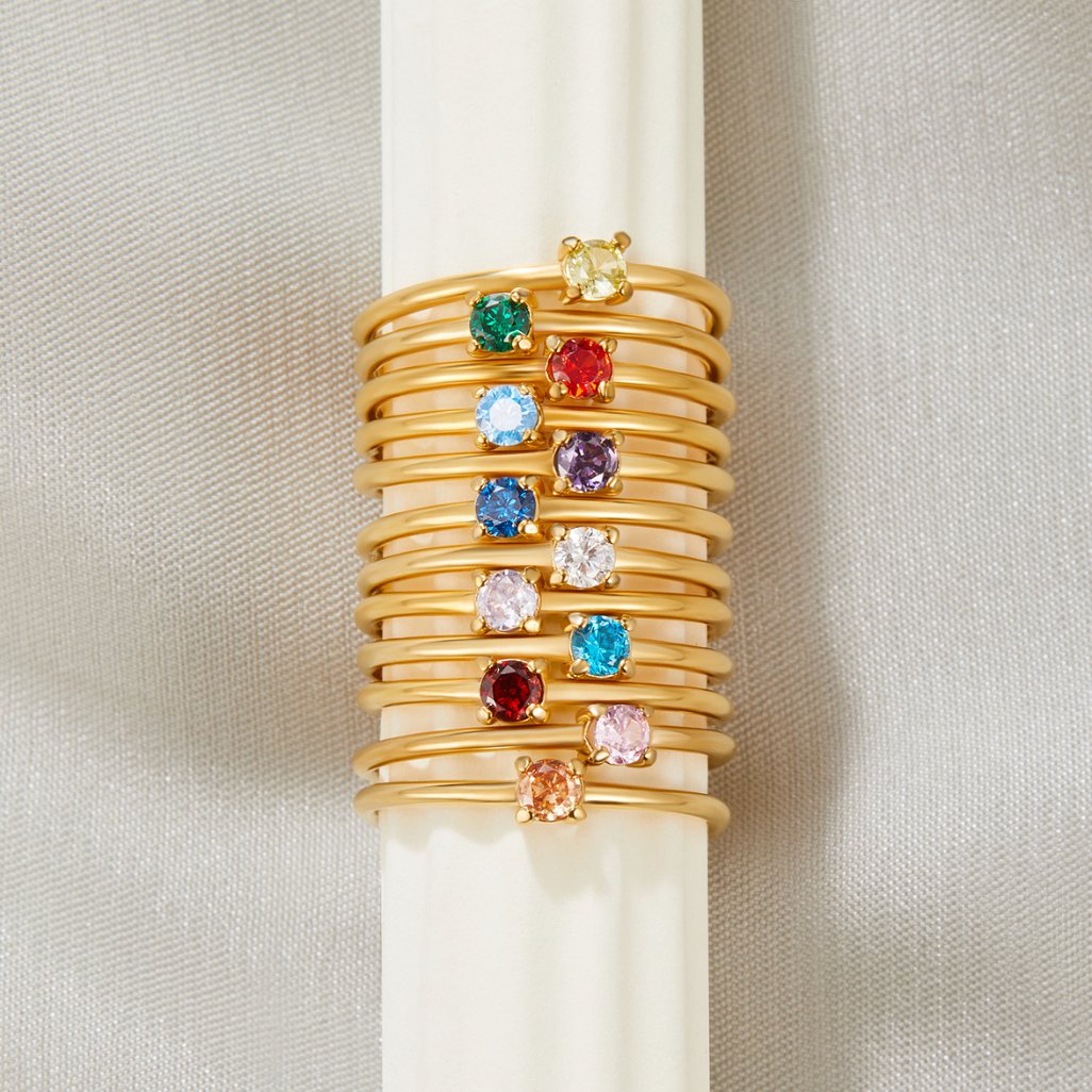 Birthstone 18K Gold Rings, stacked on a cream cylinder, twelve gold rings with coloured gemstones
