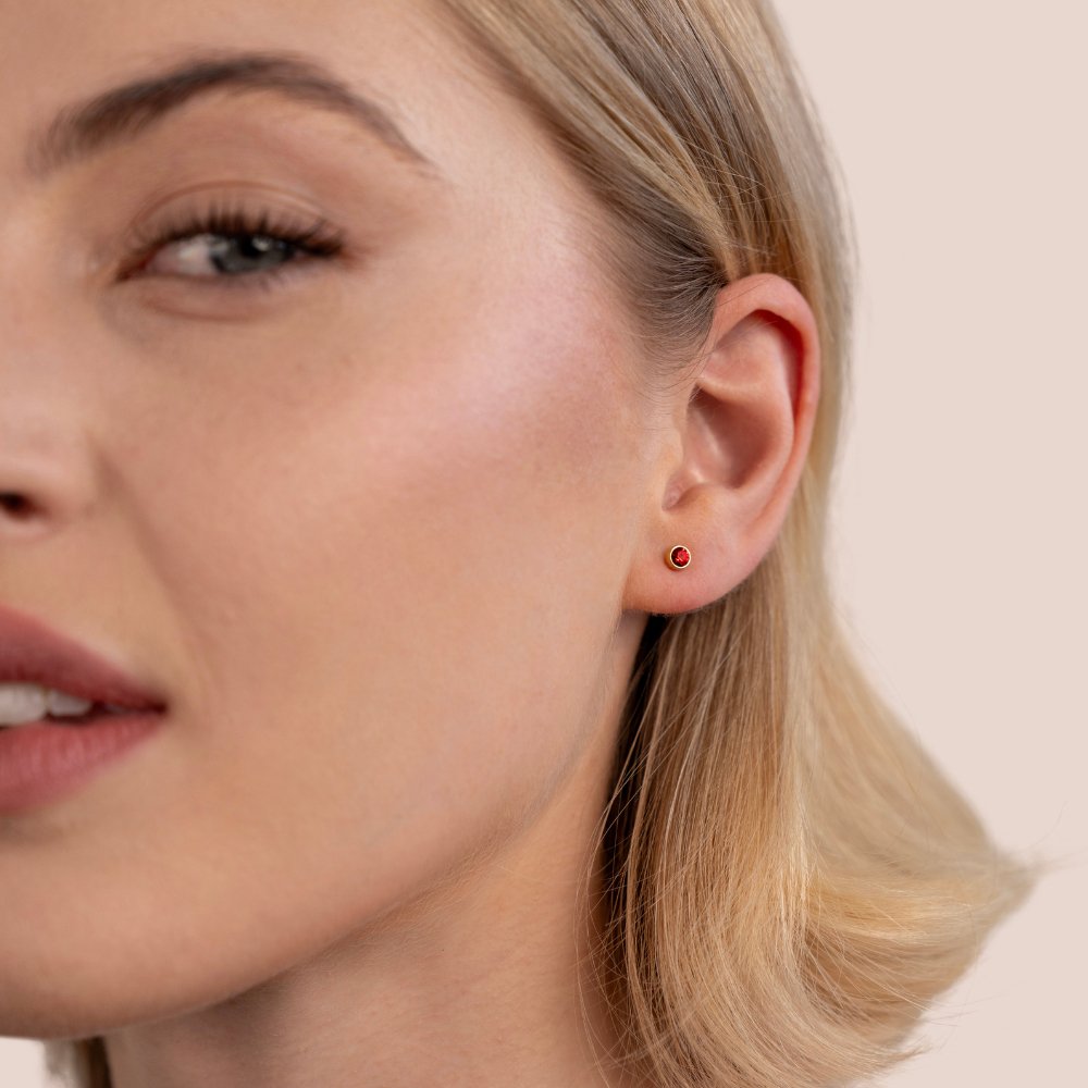 Birthstone Stud Earrings, worn on an ear, small round red stone in a gold setting