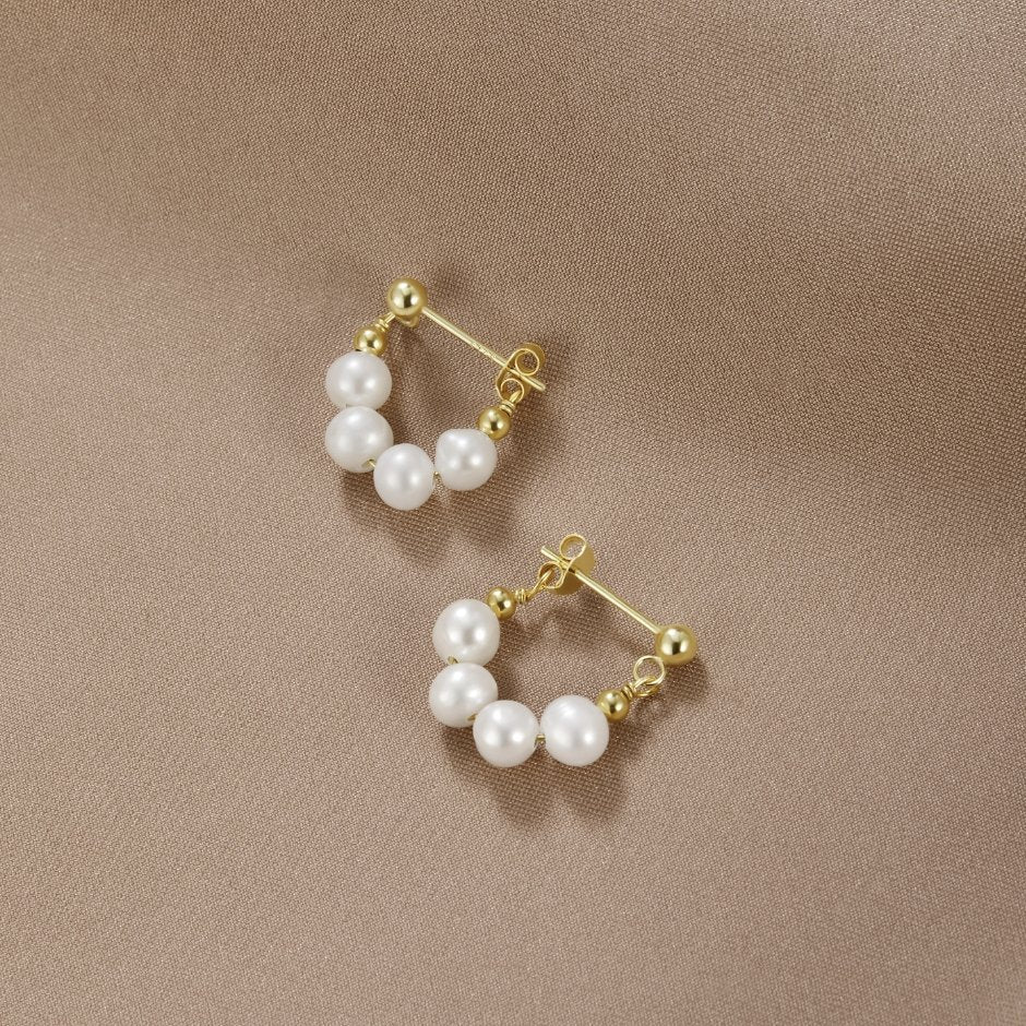 Celine Pearl Hoop Earrings, a pair of gold earrings with white pearls on beige fabric