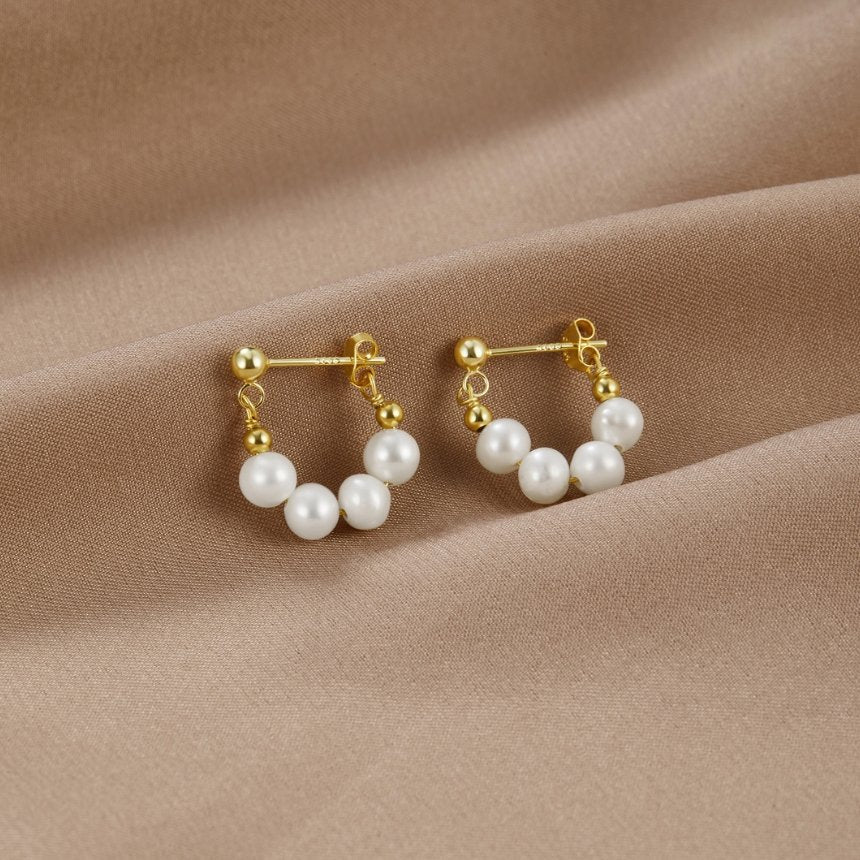 Celine Pearl Hoop Earrings, a pair of gold earrings with white pearls, lying on draped beige fabric