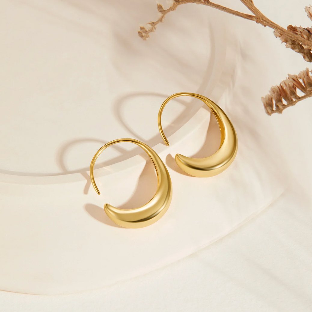 Classic Drop Hoop Earrings, two gold crescent hoop earrings on a cream surface with dried flowers