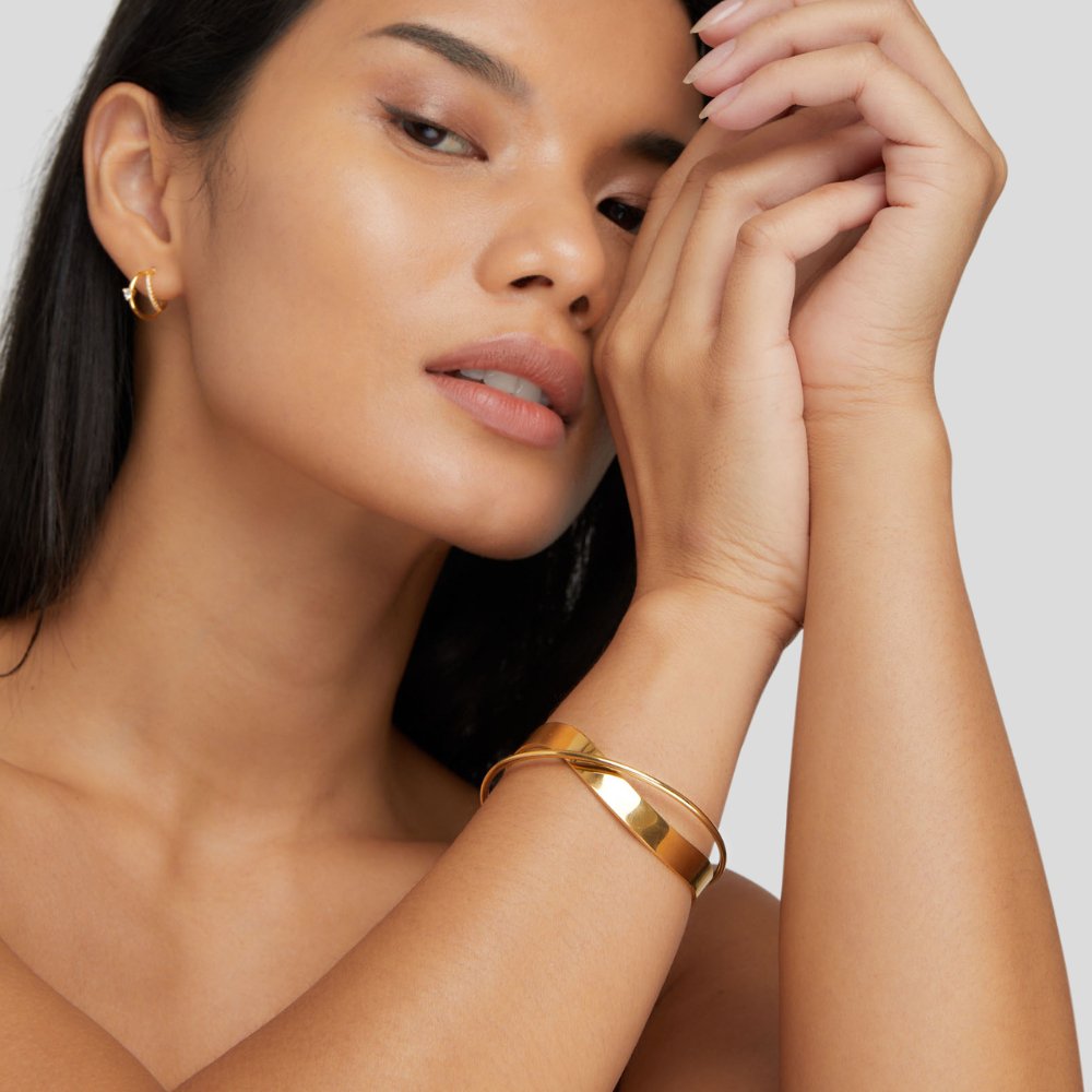 Eve Gold Bangle, worn on a wrist, with a gold hoop earring visible