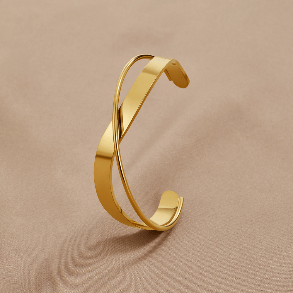 Eve Gold Bangle, open cuff lying on a beige fabric, crossed bands in polished gold
