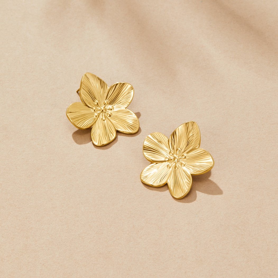 Gold Blossom Earrings, a pair of gold flower-shaped stud earrings lying on a beige surface