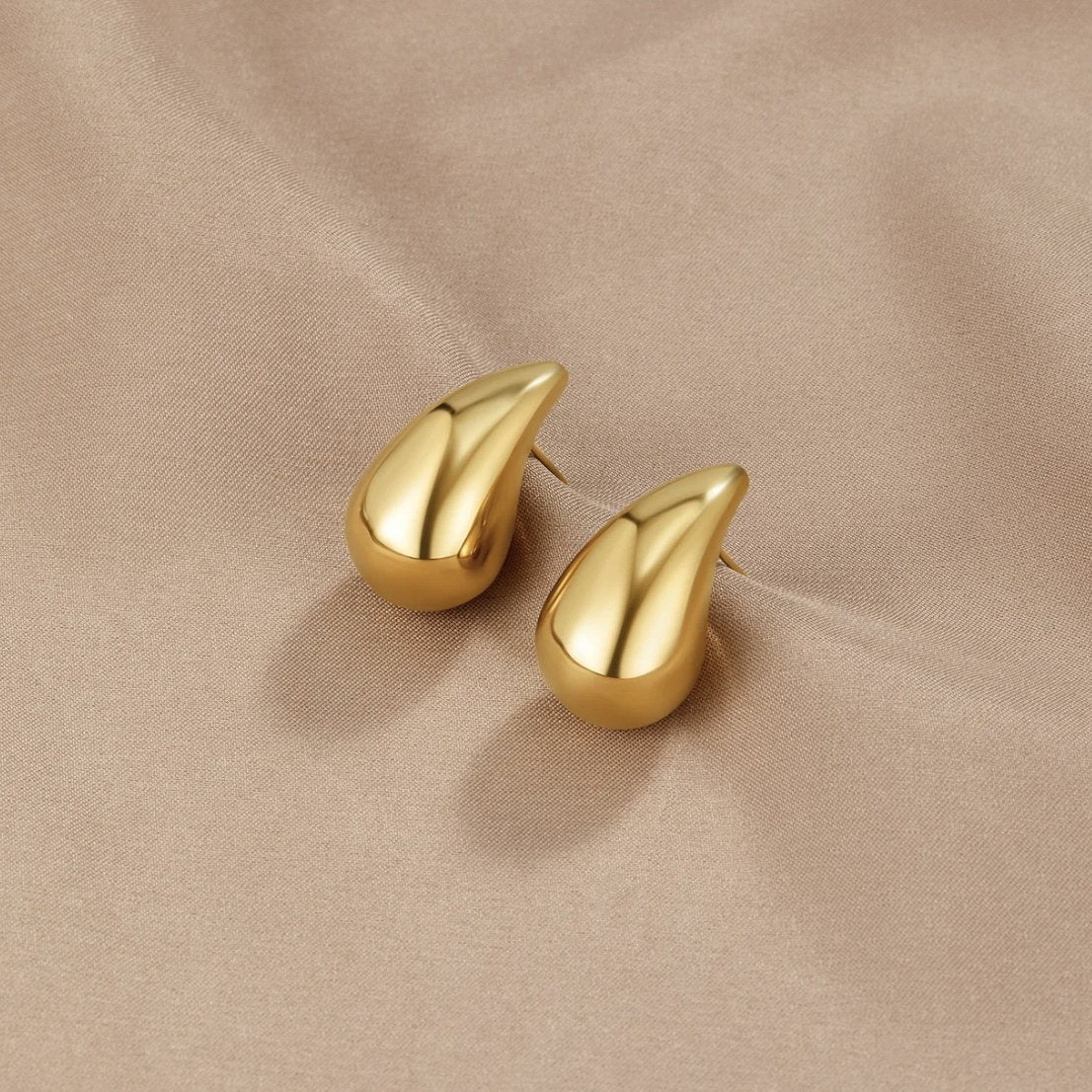 Amara Gold Teardrop Earrings, pair of polished gold teardrop earrings lying on beige satin