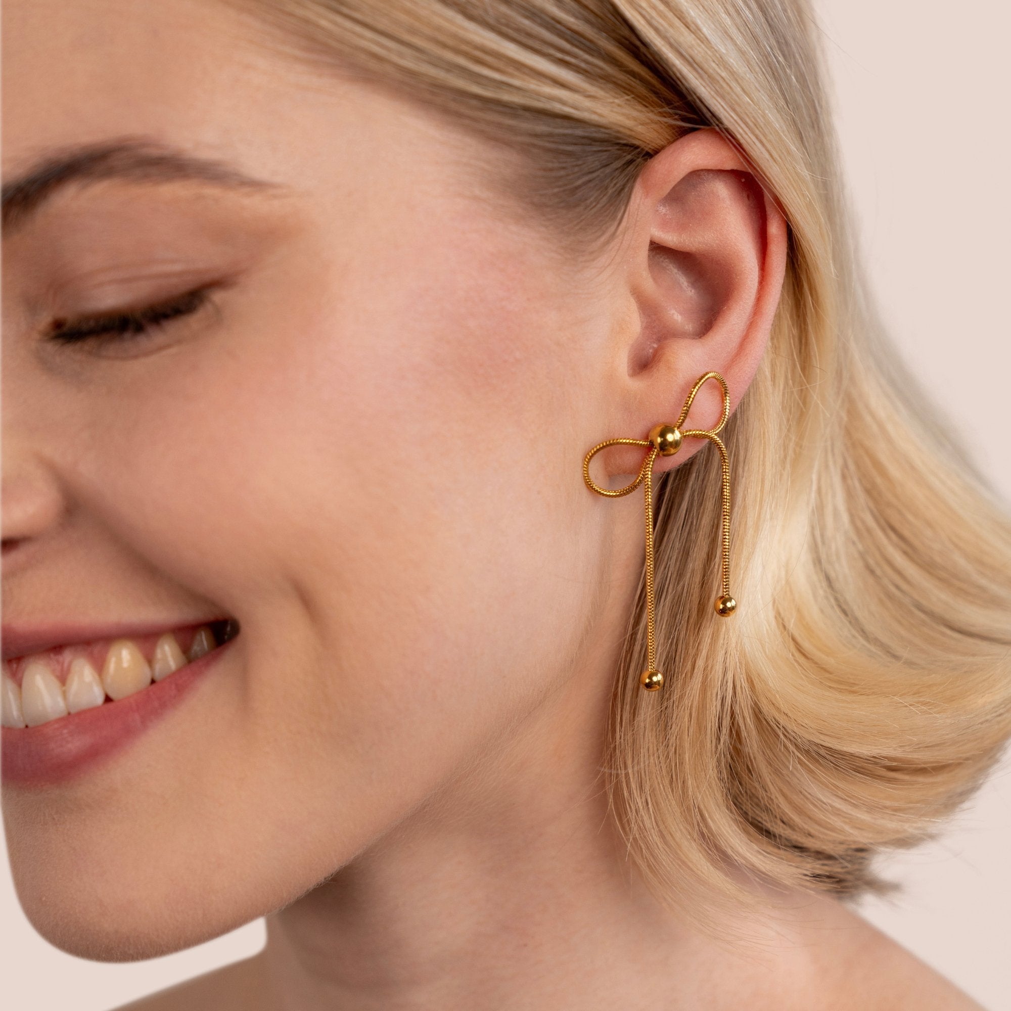 Katie Bow Earrings, worn on an ear by a smiling blonde model, gold bow with long drop