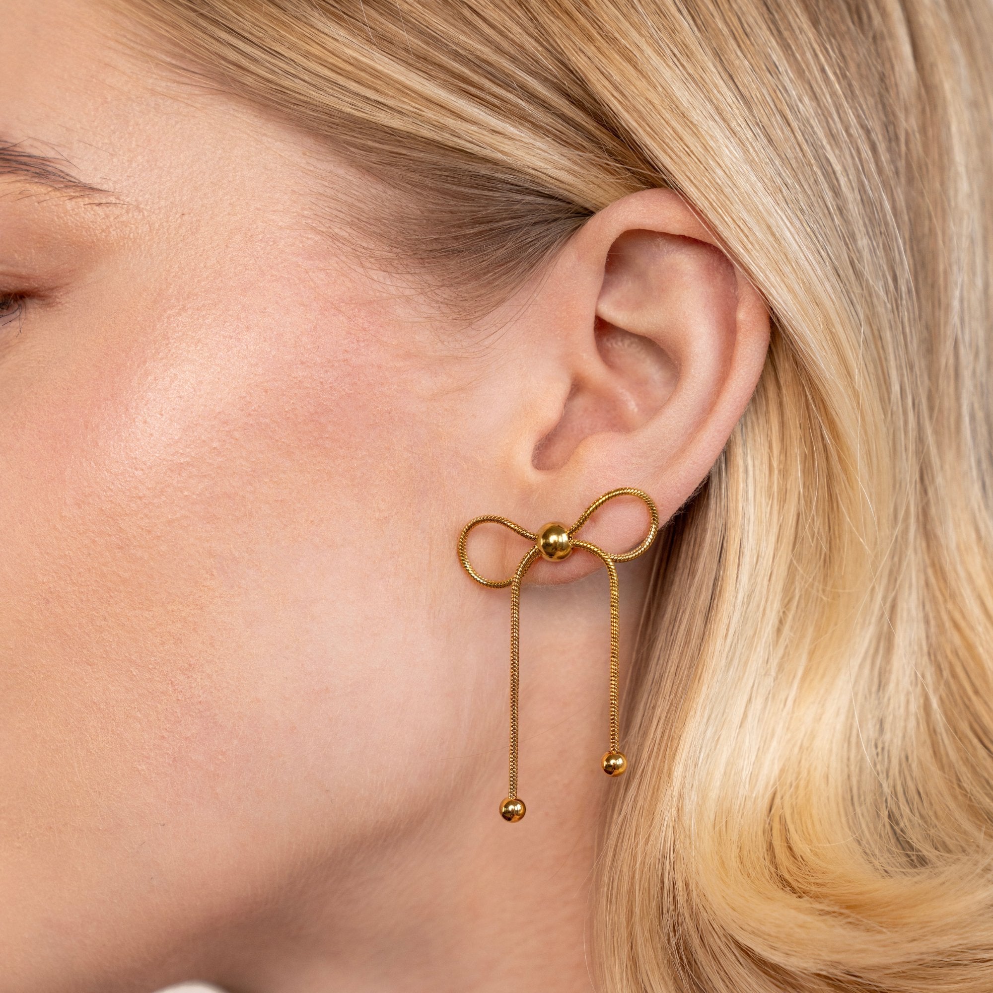 Katie Bow Earrings, worn on an ear, gold bow with two dangling ball-tipped strands