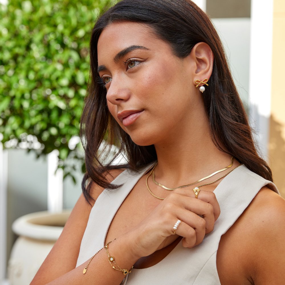 Laila Bow Pearl Earrings, worn on a model with layered gold necklaces and bracelets