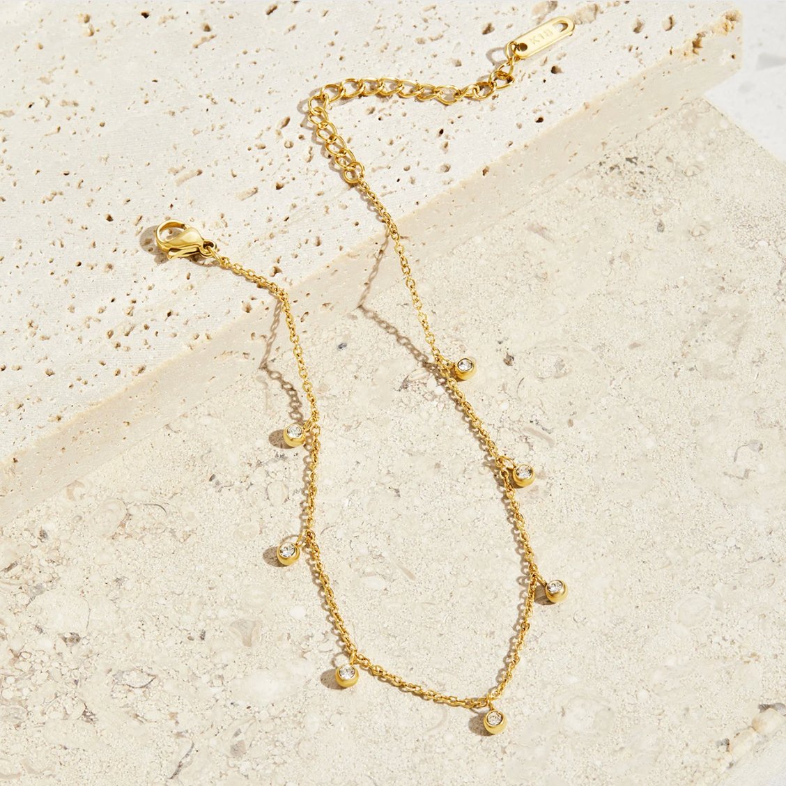 Lana Crystal Droplet Anklet, laid flat on textured stone with gold chain and small crystal drops