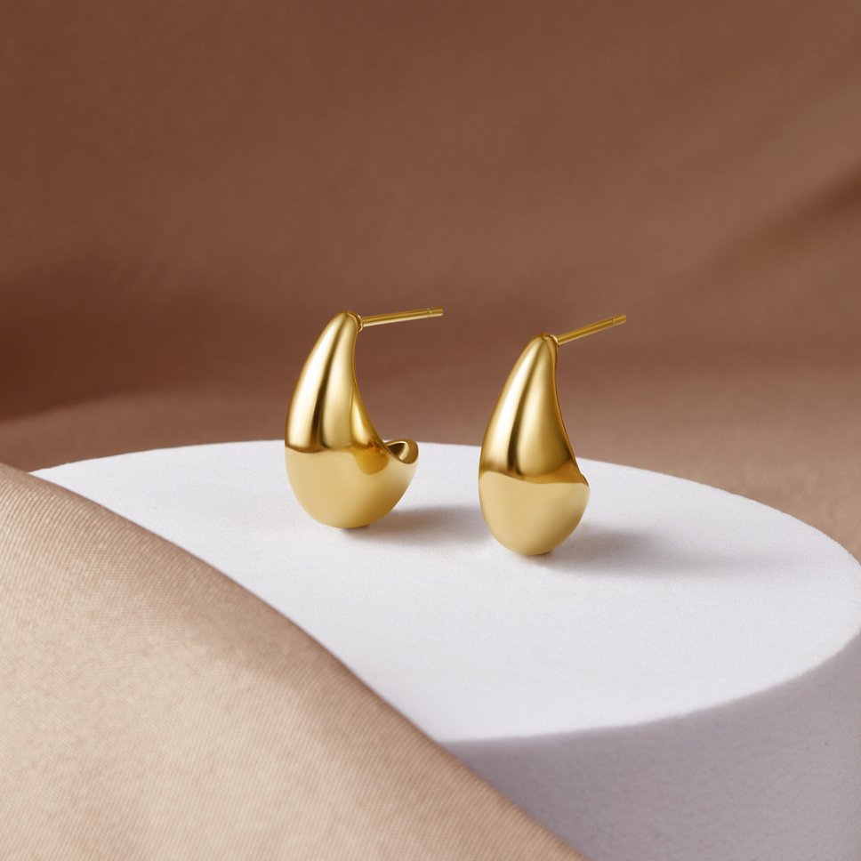 Mini Curved Gold Hoops, a pair of gold earrings resting on a white pedestal against a brown backdrop