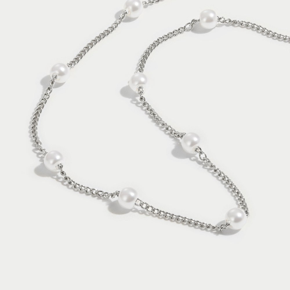 Nura Pearl Silver Necklace, silver chain necklace with white pearls, laid flat on a pale surface