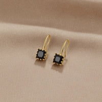 Piper Black Crystal Drop Earrings, a pair of gold earrings with square black stones on beige
