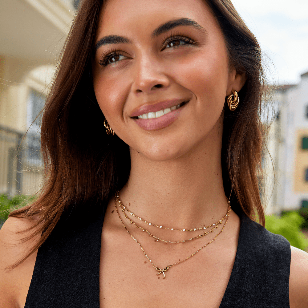 Serenity Bow Pendant Necklace, worn on a smiling model with gold hoop earrings and layered chains